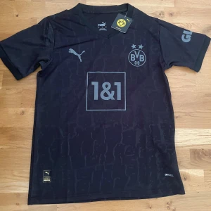 Svart Borussia Dortmund fotbollströja Puma - Snygg svart Borussia Dortmund fotbollströja från Puma med klubbmärke, sponsorlogga och diskret mönster. Tröjan har korta ärmar, V-ringning och är tillverkad i ett lätt och ventilerande material med Drycell-teknologi.
