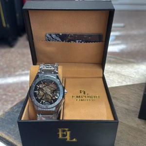 Silver automatisk klocka Emporio Limited - Snygg automatisk klocka från Emporio Limited med silverfärgat armband och boett. Urtavlan har ett öppet skelettdesign där du ser de mekaniska detaljerna i guld och svart. Klockan levereras i en lyxig svart box med gulddetaljer.