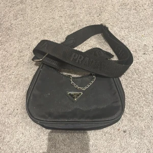 Svart axelväska med kedja - Svart axelväska  med bred axelrem där loggan är broderad. Väskan har en snygg silverfärgad kedjedetalj framtill och klassisk trekantig logga. Materialet är slitstark syntet och väskan stängs med dragkedja. Perfekt storlek för det viktigaste. Den lilla plånboken som brukar hänga på väskan borttappad (stör inte funktionen alls) därav priset❤️