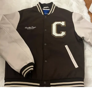 Brun och beige bomberjacka Jack & Jones - Snygg bomberjacka från Jack & Jones i storlek L. Jackan är brun med beige ärmar och detaljer, har ribbade muddar och krage med ränder, samt en stor vit C-patch på bröstet. Stängs med knappar och har två fickor framtill. Perfekt för dig som gillar college-stil.