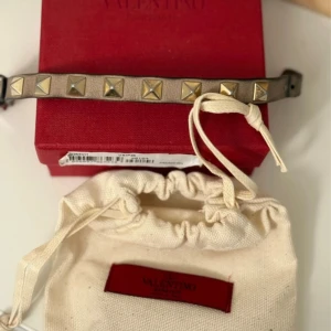 Valentino armband med nitar - Coolt armband från Valentino i beige skinn med ikoniska pyramidformade guldfärgade nitar. Justerbar längd med flera hål för perfekt passform. Kommer med original ask och dustbag. Perfekt accessoar för att lyfta din outfit.