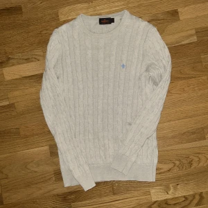 Ljusgrå kabelstickad tröja Morris - Kabelstickad ljusgrå tröja från Morris i 100% bomull. Klassisk rund halsringning och diskret blå brodyr på bröstet. Perfekt för dig som gillar stilrena och tidlösa plagg med snygg struktur. Långärmad och ribbade muddar för extra komfort.