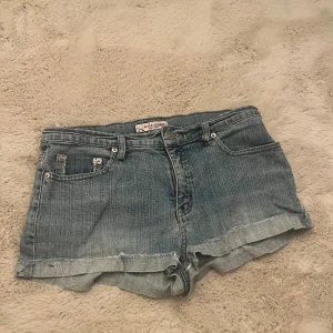 Blå jeansshorts från M.A.P.P - Snygga blå jeansshorts från M.A.P.P med klassisk femficksdesign och uppvikta benslut. Shortsen har normal passform och är tillverkade i bomull. Perfekta för sommaren och har en avslappnad vibe med slitna detaljer.
