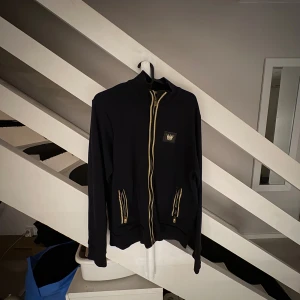 MR Originals Zip Mörkblå - Svart hoodie från MR original smed guldfärgade dragkedjor och detaljer. Tröjan har lång ärm, två fickor framtill och en broderad krona på bröstet samt i nacken. Perfekt för dig som gillar streetstyle och snygga detaljer.