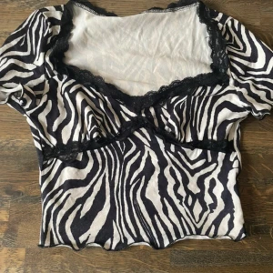 Zebramönstrad topp med spetsdetaljer - Säljer en kortärmad topp med svartvitt zebramönster och snygga svarta spetsdetaljer längs v-ringningen och under bysten. Toppen har vågig nederkant och är croppad för en trendig look. Perfekt för dig som vill sticka ut med ett djurmönster.