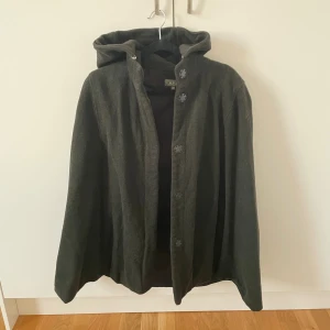 Mörkgrön poncho med huva från A.P.C. - Stilren mörkgrön poncho från A.P.C. i ull med huva och knäppning framtill. Klassisk och enkel design med rak passform. Kappan har stora knappar och en mjuk känsla. 
