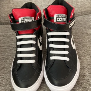 Converse Cons svarta och röda sneakers - Säljer ett par Converse Cons sneakers i svart och rött skinn med vita detaljer och snörning. Skorna har högt skaft, kardborreband och klassisk Converse-stjärna på sidan. Snygg kontrast mellan färgerna och cool streetstil. Perfekta för dig som gillar sportig vibe.