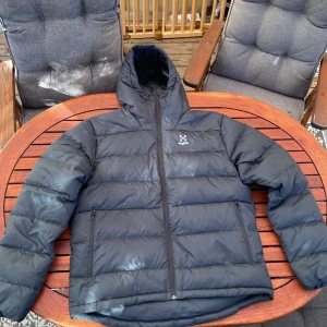 Svart dunjacka från Haglöfs XL (Bield down Hood) - Svart dunjacka från Haglöfs i storlek XL, modell Bield down Hood. Jackan har huva, två dragkedjeförsedda fickor och quiltad design. Perfekt för kalla dagar med sin fluffiga och varma känsla. Klassisk look med diskret logga på bröstet och hel dragkedja framtill. I mycket fint skick, knappt använd överhuvudtaget.