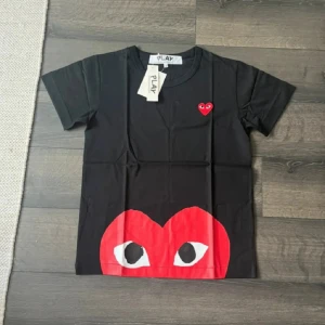 CDG T-shirt - Helt ny Svart t-shirt från Comme des Garçons Play med ikoniskt rött hjärta med ögon på bröstet och stor röd hjärtdetalj med ögon längst ner. Klassisk rund hals och korta ärmar. Tillverkad i mjuk bomull för en skön känsla.