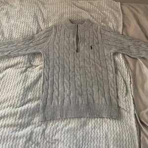 Grå kabelstickad tröja Polo Ralph Lauren - En helt ny Kabelstickad grå tröja från Polo Ralph Lauren med halv dragkedja och ribbade muddar. Klassisk design med broderad logga på bröstet och hög krage. Perfekt för lager-på-lager under kyliga dagar.