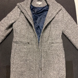 Grå ullmix kappa från H&M - Säljer en grå kappa från H&M i ullmix med snygg struktur och rak passform. Jackan har dragkedja framtill och två fickor med dragkedja. Perfekt för dig som gillar stilrena och klassiska ytterplagg med modern touch.