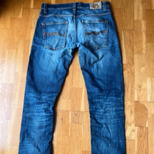 Blå jeans från Nudie Jeans, 32/34 - Säljer ett par klassiska blå jeans från Nudie Jeans i storlek 32/34. Jeansen har snygga slitningar, kontrastsömmar och Nudie-logga på bakfickorna. Modellen har rak passform och är tillverkad i robust denim med fem fickor. Ett litet hål finns i skrevet och i högra fickan. Nudie har gratis reparationer av deras jeans👍🏼Model Grim Tim