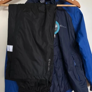 Blå vind set från Trekmates - Säljer ett set Trekmates med huva och dragkedja o byxor. Jackan har två fickor med dragkedja och justerbara muddar. Perfekt för outdoor-äventyr