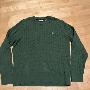 Grön kabelstickad tröja från Gant - Mörkgrön kabelstickad tröja från Gant med rund halsringning och broderad logga på bröstet. Tröjan har långa ärmar och ribbade muddar vid ärmslut och nederkant. Perfekt för lager-på-lager och snygg till jeans.