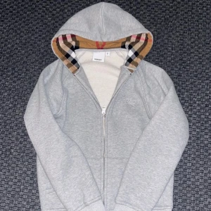 Grå hoodie från Burberry med rutig huva - Snygg grå hoodie från Burberry med dragkedja och klassiskt broderat logotypmärke på bröstet. Huvan är fodrad med ikoniskt rutigt Burberry-tyg i beige, rött och svart. Perfekt för dig som vill ha en stilren och exklusiv look. Priset kan diskuteras!