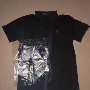 Svart pikétröja  - Svart pikétröja med klassisk krage och knappar framtill. Ikonisk röd broderad logga på bröstet. 