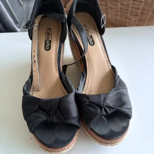 Svarta kilklack-sandaler från Din Sko - Snygga svarta sandaler med kilklack från Din Sko. Skorna har öppen tå, rosett framtill och justerbart spänne vid ankeln. Ovandelen är i tyg och kilklacken är klädd i flätad jute för en cool sommarkänsla. Perfekt för dig som gillar att sticka ut med stil.