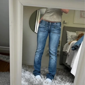 Blå lågmidjade jeans med raka ben - Säljer ett par blå jeans med låg midja och raka ben. Jeansen har snygga slitningar, dubbla bälteshällor och stora framfickor med coola detaljer. Materialet är klassisk denim och passformen är normal. Perfekta för dig som gillar en avslappnad men trendig look.
