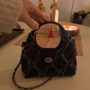 Mörkblå axelväska med kedja - Snygg mörkblå axelväska(jeans Matrial) med coolt rutigt mönster i fransig denim och silverfärgad kedja. Väskan har ett runt handtag och ett dekorativt spänne framtill. Perfekt för dig som gillar unika detaljer och vill sticka ut.
