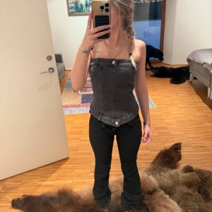 Svart denimkorsett med dragkedja  - Säljer en svart korsett-topp i denim med markerade sömmar och bälteshällor. Toppen har smockad rygg, dragkedja bak och knappdetaljer framtill. Axelbanden är justerbara och modellen är croppad med en tight passform. Perfekt för att lyfta vilken outfit som helst. 