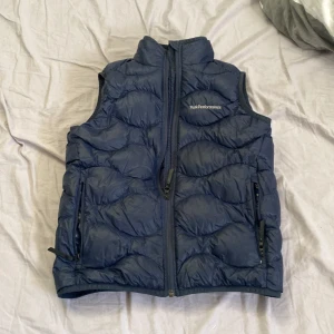 Blå dunväst från Peak Performance - Snygg blå dunväst från Peak Performance med quiltad design och hög krage. Ger en rikigt fräsch och clean look den är i storlek 15 men passar xs. Det enda negativa är en liten fläck i insidan men syns ej när den är på. 