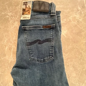 Nudie Jeans blå jeans W28 L32 HELT NYA! - Säljer ett par klassiska blå jeans från Nudie Jeans i modellen W28 L32. Jeansen har fem fickor, raka ben och snygga slitningar framtill. Tillverkade i ekologisk bomull och har Nudie-loggan på bakfickan. Perfekta för dig som gillar stilrena och tidlösa jeans.