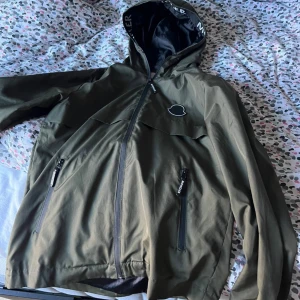 Olivgrön vindjacka från Moncler - Snygg olivgrön vindjacka från Moncler med huva och svarta detaljer. Skriv vid minsta lilla fråga. S