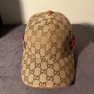 Gucci keps med GG-mönster och ränder - Exklusiv keps från Gucci i beige och brunt med klassiskt GG-monogram över hela ytan. Sidorna har gröna och röda ränder för den ikoniska Gucci-looken. Kepsen är tillverkad i bomull och har detaljer i läder. Perfekt accessoar för att sticka ut.