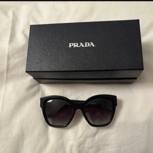 Svarta cat eye-solglasögon från Prada - Snygga svarta solglasögon från Prada med cat eye-form och mörka glas. Kommer med originaletui i svart med Pradas logga. Perfekta för dig som vill ha en lyxig och trendig accessoar till din outfit.