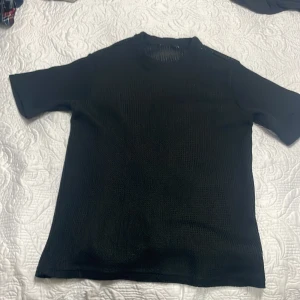 Svart stickad t-shirt med struktur - Svart t-shirt med korta ärmar i ett stickat material som ger en cool, strukturerad look. Modellen har rund hals och är lätt transparent tack vare den grova stickningen. Passar dig som gillar en unik och edgy stil.