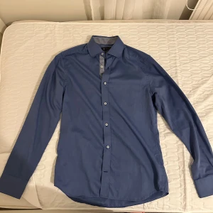 Blå slim fit skjorta från Riley - Snygg blå skjorta från Riley i slim fit-modell. Skjortan har långa ärmar, vita knappar och ett rutigt mönster på insidan av kragen och knappslån. Perfekt för dig som gillar stilrena och klassiska plagg.