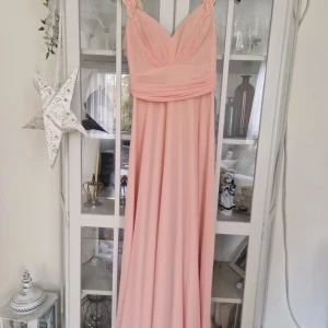 Ljusrosa långklänning med rosett - Elegant ljusrosa långklänning med v-ringad omlottdesign och breda axelband med broderade detaljer. Klänningen har en stor rosett och volang baktill samt markerad midja. Tillverkad i ett lätt, flytande material som ger ett mjukt fall. Rosa sjal som tillbehör 
