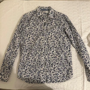 Blommig skjorta från Premium - Vit skjorta med mörkblått blommönster från Premium. Skjortan har klassisk krage, knäppning framtill och långa ärmar. Tillverkad i mjuk bomull, perfekt för dig som gillar att sticka ut med snygga detaljer.