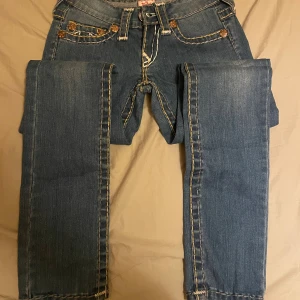 True Religion jeans med kontrastsömmar - Säljer ett par blå jeans från True Religion med ikoniska kontrastsömmar i vitt och gult. Byxorna har raka ben, klassiska fickor och tydlig logga bak. Snygga detaljer med grova sömmar och broderade bakfickor som sticker ut. Står 27 på byxorna men skulle säga de är 24-25. Säljer på grund av att de är för små 💕