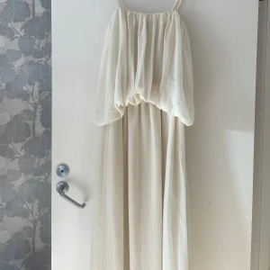 Klänning - Helt underbar smörvit  långklänning med omlott och volangdetaljer. Klänningen har tunna axelband och ett luftigt, flowy tyg som ger en romantisk vibe. Perfekt för sommarens alla tillfällen. Passar dig som gillar boho!