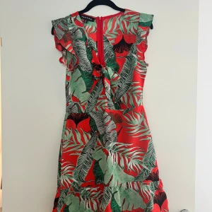 Röd blommig kortklänning från Parisian/Boohoo - Snygg röd kortklänning från Boohoo med tropiskt blommigt mönster i grönt, svart och vitt. Klänningen har volangdetaljer vid axlarna, knyt framtill och är ärmlös. Perfekt för sommarens alla tillfällen när du vill sticka ut med färg och mönster.