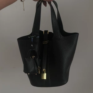 Svart bucket handväska med hänglås - Stilren svart bucket handväska i skinn med grov struktur och guldfärgade detaljer. Väskan har två rejäla handtag, dekorativt hänglås och en miniatyrväska som accessoar. Perfekt för dig som gillar minimalistisk och lyxig vibe.
