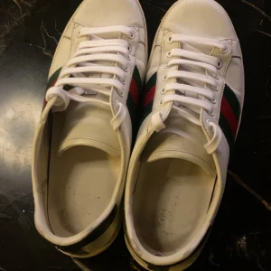 Gucci vita sneakers  - Säljer ett par klassiska vita sneakers från Gucci med de ikoniska röd-gröna ränderna på sidorna. Skorna har rund tå, platt sula och snörning. Tillverkade i skinn och har en stilren, tidlös look som passar till det mesta.