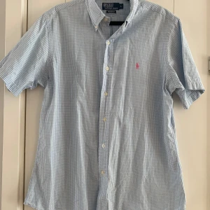 Blårutig kortärmad skjorta Polo Ralph Lauren - Kortärmad skjorta från Polo Ralph Lauren i custom fit. Skjortan är blårutig med små vita rutor, har klassisk krage, knappar framtill och en rosa broderad logga på bröstet. Tillverkad i tunn bomull, perfekt för varma dagar. Mått Över bröstet: ca 58 cm Längd från axeln och ner: ca 75 cm  Material: 100% bomull  Kommer från ett djur- och rökfritt hem. 
