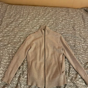 Beige kofta från Massimo Dutti - Säljer en stilren beige kofta från Massimo Dutti i mjuk ull- och kashmirblandning. Koftan har hel dragkedja framtill, ribbade muddar och hög krage. Perfekt för lager-på-lager och passar till allt i garderoben.