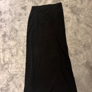 Svart långkjol i mjukt tyg - En stilren svart långkjol med rak passform. Kjolen är enkel och minimalistisk, perfekt att matcha med olika toppar. Tillverkad i ett mjukt och följsamt material som ger en snygg siluett.