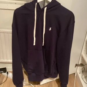 Mörkblå hoodie från Polo Ralph Lauren - Säljer en mörkblå hoodie från Polo Ralph Lauren med vit broderad logga på bröstet och vita snören i huvan. Klassisk känguruficka framtill och ribbade muddar. Perfekt för dig som gillar stilrena och bekväma plagg.
