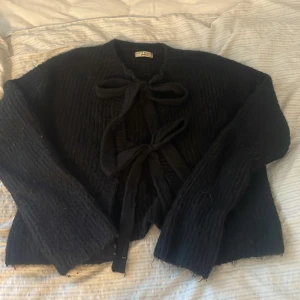 Svart stickad kofta med knyt från Gina Tricot - Mysig svart stickad kofta från Gina med två stora knytband framtill. Koftan har lång ärm, rak passform och grovstickad struktur. Använt få gånger!!