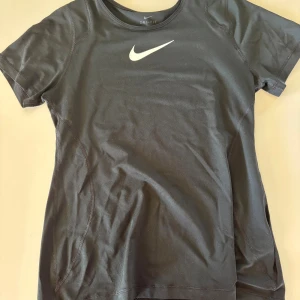 Svart Nike Dri-Fit t-shirt  - Svart Nike Dri-Fit t-shirt i funktionsmaterial med klassisk vit swoosh-logga på bröstet. T-shirten har rund halsringning och korta ärmar, perfekt för dig som gillar sportig och clean stil.