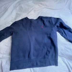 Mörkblå sweatshirt från Polo Ralph Lauren - Bra skick, knappt använd, snygg. Klassisk mörkblå sweatshirt från Polo Ralph Lauren med rund hals och diskret röd logga broderad på bröstet. Tröjan har långa ärmar och är tillverkad i mjuk bomullsmix, perfekt för en chill och stilren look.