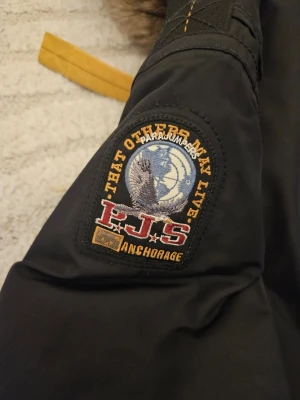 Svart Parajumpers parkas med päls - Snygg svart parkajacka från Parajumpers med pälsfodrad huva och gul detalj vid halsen. Jackan har flera fickor framtill, dragkedja och knappar. Perfekt för kalla dagar med sin varma och robusta design. Jackan behöver tvättas har inte använt den på länge.