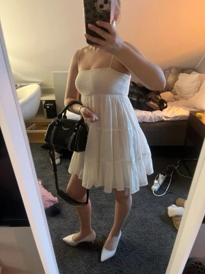 Vit kort sommarklänning med tunna band - Söt vit kortklänning med tunna axelband och smockad överdel. Klänningen har volangdetaljer och en luftig, flowy passform som passar perfekt till sommaren. Tillverkad i ett lätt material som känns bekvämt och fräscht. Man kan ha den som kjol eller klänning. Lappen är bortklippt! Den har några defekter men inget man ser vid användning