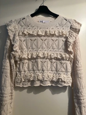 Vit stickad volangtröja från Zara - Supersöt vit stickad tröja från Zara med volanger och spetsdetaljer framtill. Tröjan har lång ärm, rund halsringning och ett fint hålmönster i garnet. Perfekt för dig som gillar romantisk och trendig stil.