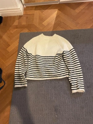 Vit stickad tröja med svarta ränder - Säljer en stickad tröja i vitt med svarta horisontella ränder på nedre delen och ärmarna. Tröjan har en avslappnad passform, rund halsringning och långa ärmar. Perfekt för en chill och trendig look.