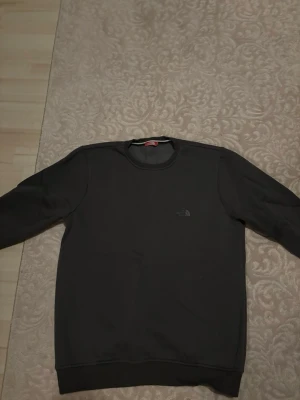 Svart sweatshirt från The North Face - En stilren svart sweatshirt från The North Face med rund halsringning och diskret logga på bröstet. Tröjan har långa ärmar och ribbade muddar vid ärmslut och nederkant. Perfekt för en avslappnad och trendig look.